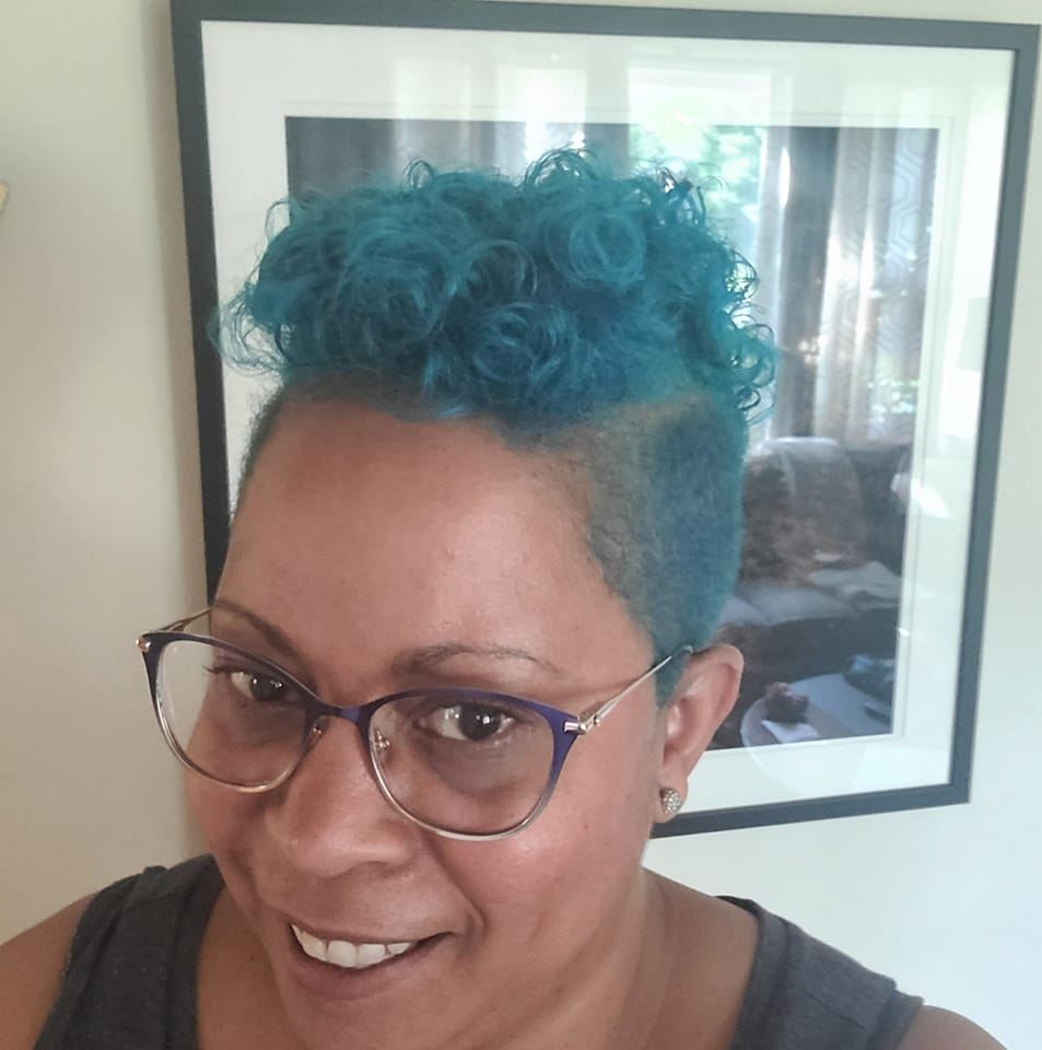 A photo of a young African American woman with large, rounded, purple framed glasses and dyed blue hair taken from a slightly elevated angle. She is smiling at the camera
