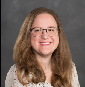 A young white woman with brown eyes, round cheeks, wire-framed glasses, and long brown hair.
